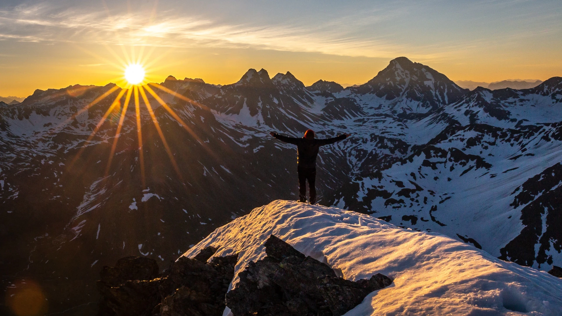 A person with their back against the camera standing with their arms stretched out to the sides, taking in a sunset from a mountain top.