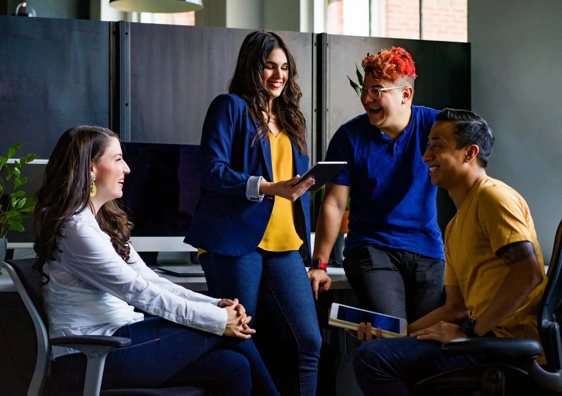 Four smiling and laughing people by a desk in an office. Two are leaning on the desk, the other two are sitting in office chairs in the foreground.