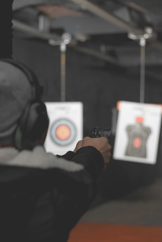 Person aiming for a target in an indoors shooting range. The focus is on the person, who is viewed from behind, with safety earmuffs, a black jacket and a gray beanie.